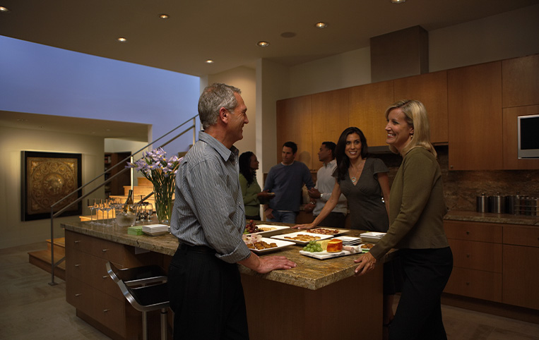 Persons gathering around an island in a modern kitchen with lights dimmed.